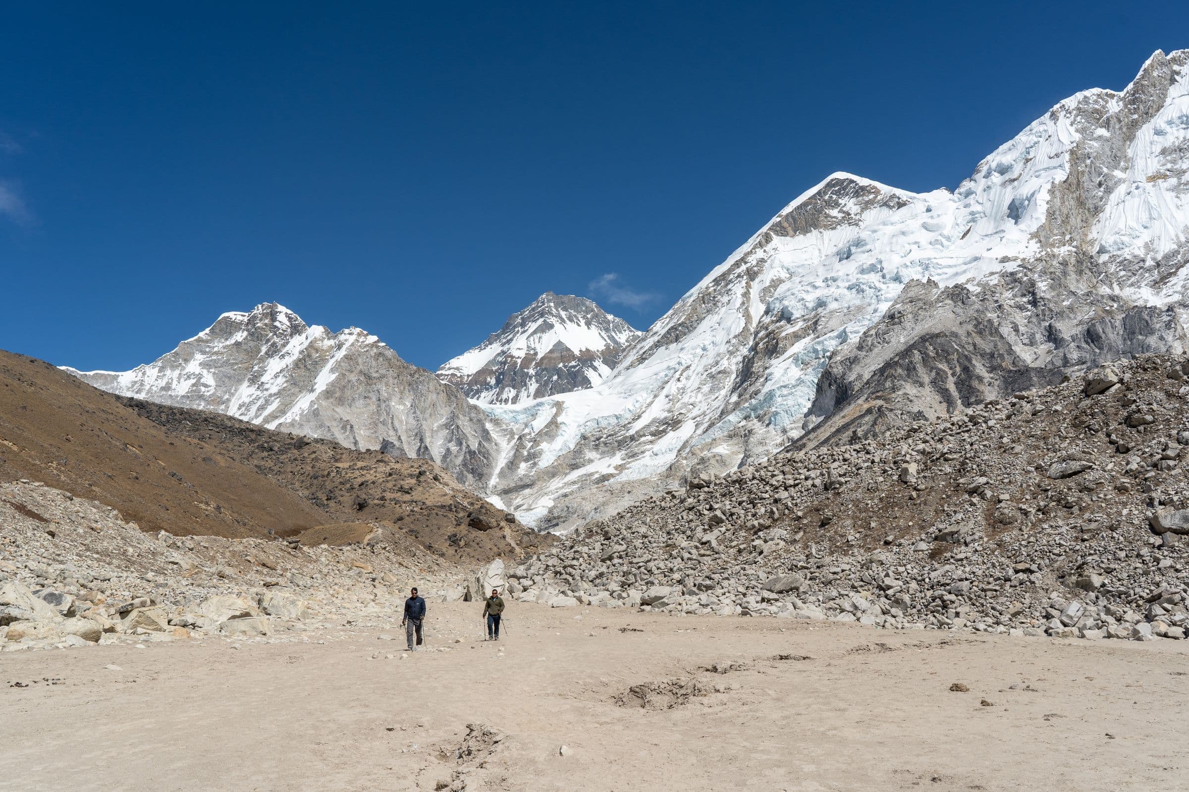 Two hikers walk towards rocky Himalayan mountains under clear blue sky.