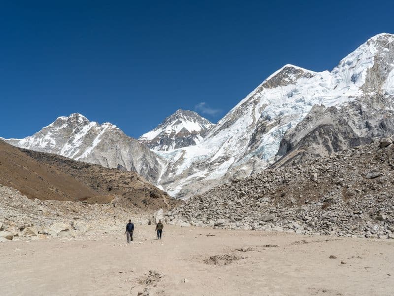 Two hikers walk towards rocky Himalayan mountains under clear blue sky.